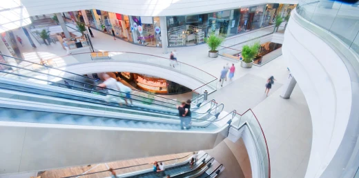 Interior of a bright shopping mall with escalators, people, plants, and storefronts.