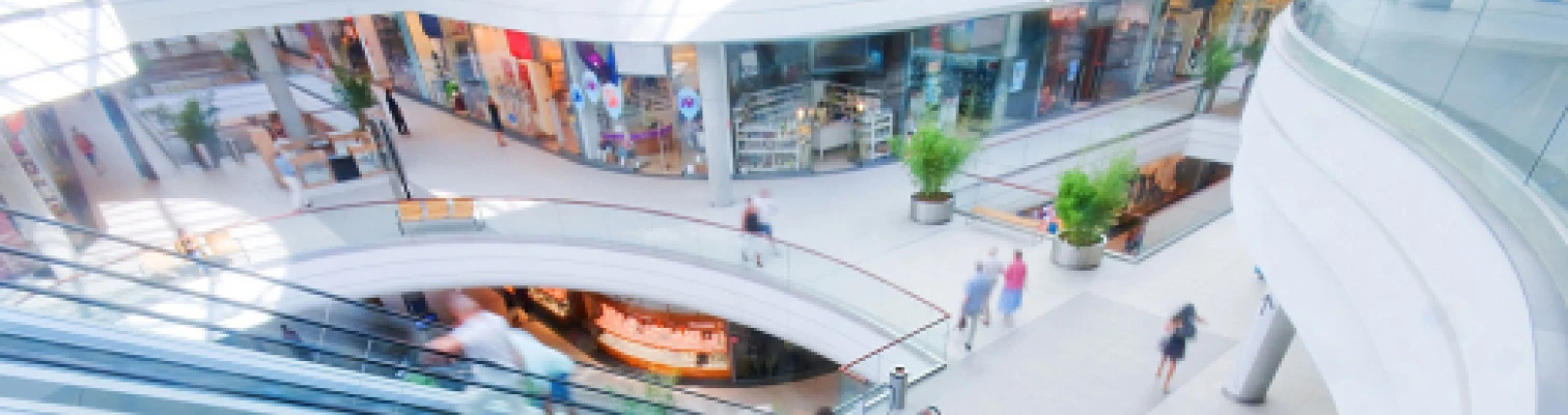 Interior of a modern shopping mall with escalators, shops, plants, and people walking.