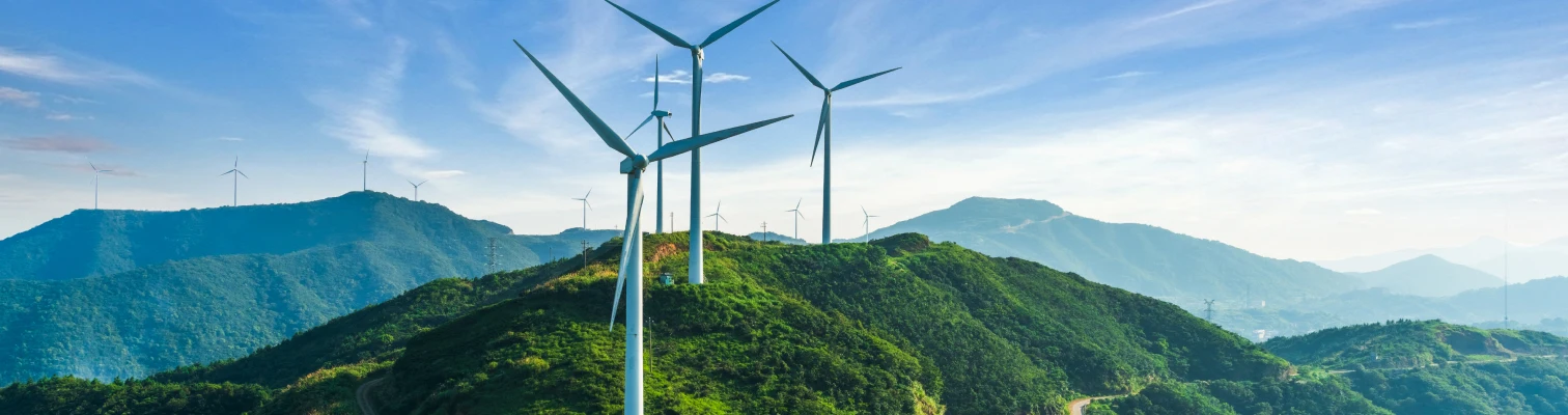 Wind turbines on green hills under a blue sky with mountains in the background.