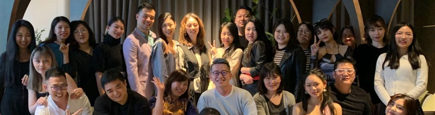 Group portrait of smiling young adults posing indoors against a curtain and wooden decor.
