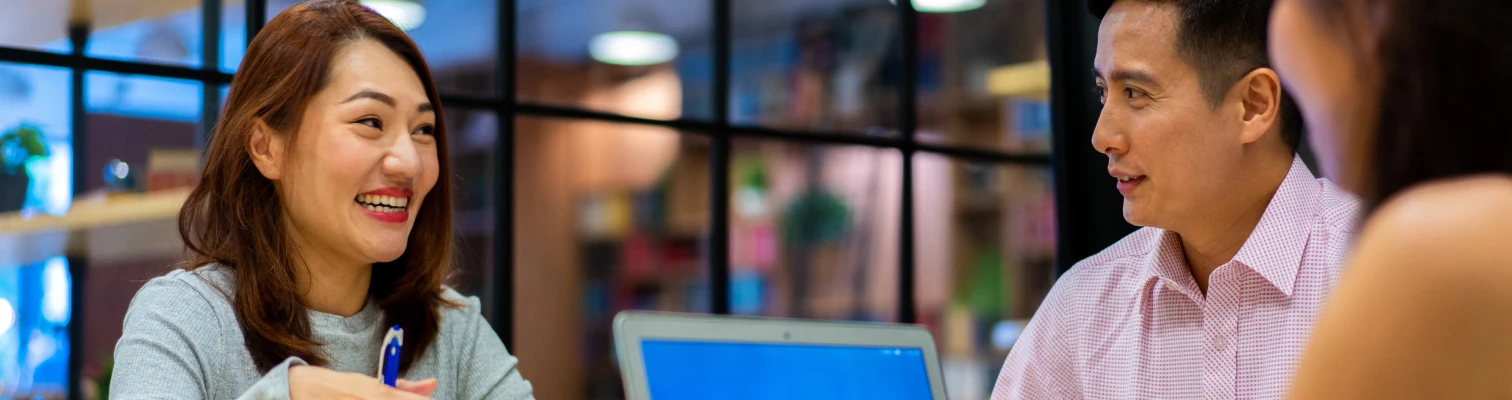 Three people smiling and talking around a laptop in a bright modern office setting.