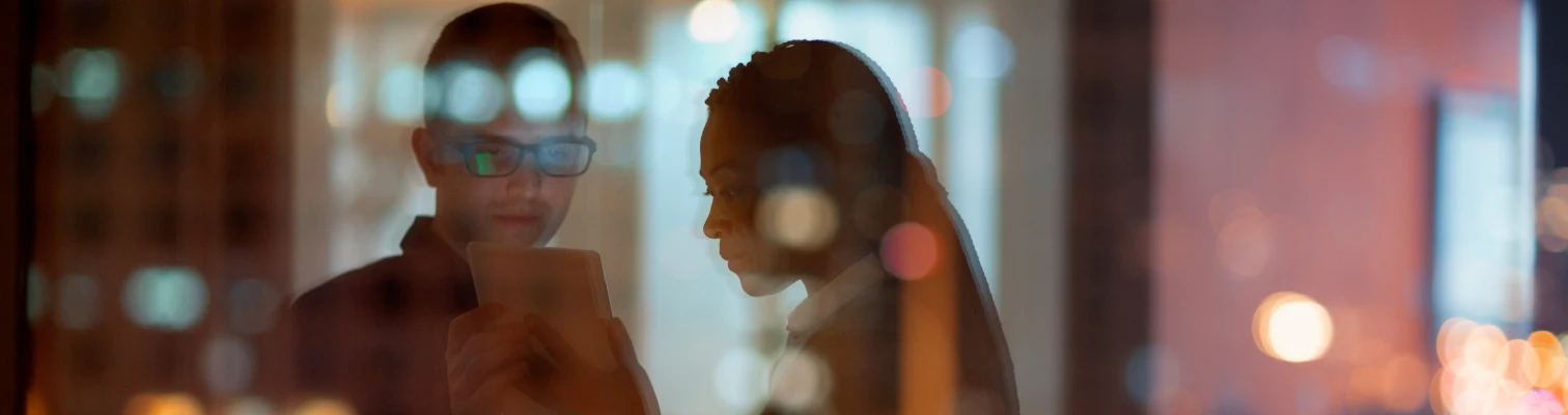 Two people indoors, seen through reflective glass, looking at a tablet or phone together.
