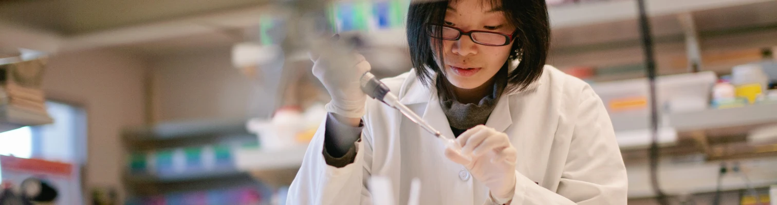 Scientist wearing glasses and a white lab coat using a pipette in a laboratory setting.